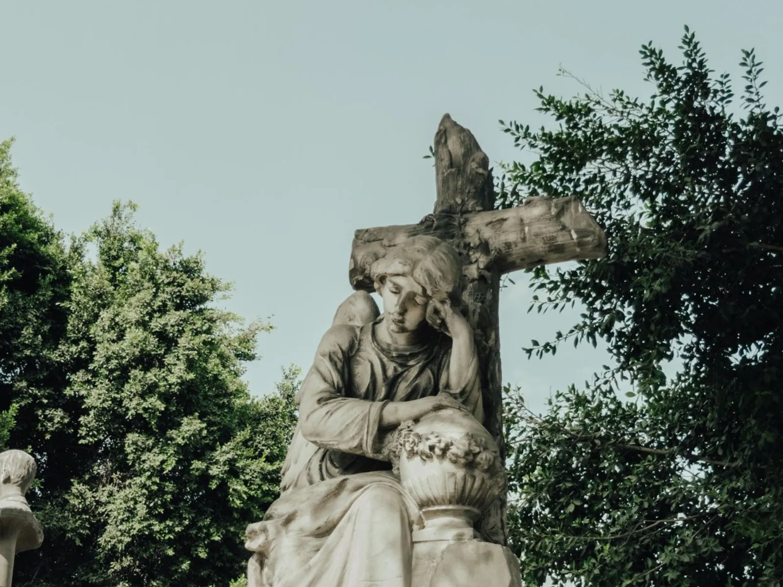 A weathered stone angel statue resting against a cross in a cemetery, surrounded by trees.